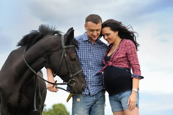 A walk on the ranch. Young couple - she is a brunette with long hair, pregnant; he is tall and brave, holding the reins of the black horse