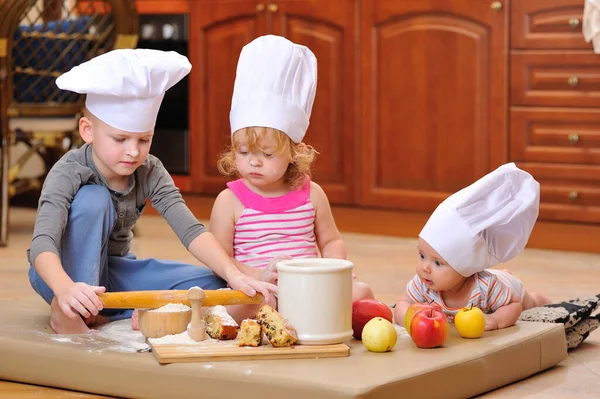 siblings - boys and girl - in chef\'s hats near the fireplace sitting on the kitchen floor soiled with flour, playing with food, making mess and having fun