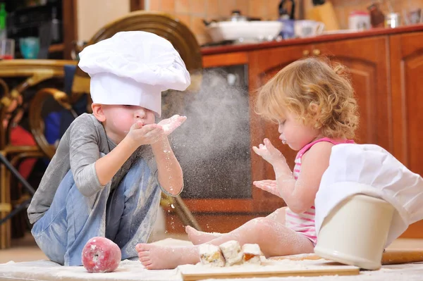 两个兄弟姐妹 男孩和女孩 戴着厨师帽 坐在厨房地板上 沾满了面粉 玩着食物 弄得乱七八糟 玩得很开心 图库图片