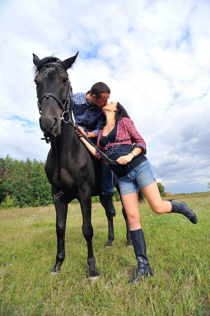 Awaiting the child, walking on the meadow. Young couple - she is a handsome brunette with long hair, pregnant; he is tall and brave, astride a black horse.
