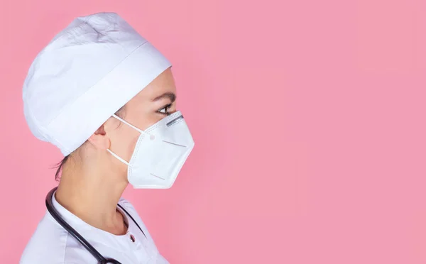 Close-up profile portrait of a young female doctor in a medical cap and protective mask on a pink background with space for text. Female doctor in the clinic. Coronavirus epidemic pandemic, covid-19