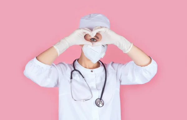 Beautiful positive face of a doctor or nurse in a uniform and medical mask. Hands in sterile gloves holding the heart shape next to the eye. Healthy heart. World humanitarian day. Isolate. Copy space