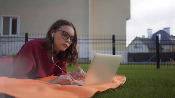 Kulaklıklı, dizüstü bilgisayarlı, çimenlikte çalışan gözlüklü kız. — Stok fotoğraf