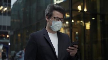 Maskeli ve telefonlu bir adam şehrin cam binalarının arka planında yürüyor.