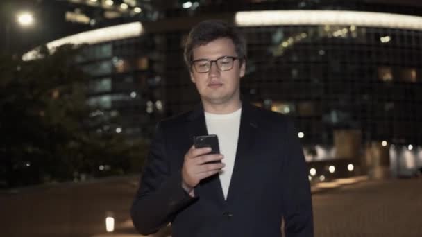 Empresário em óculos andando com o telefone no fundo de edifícios — Vídeo de Stock