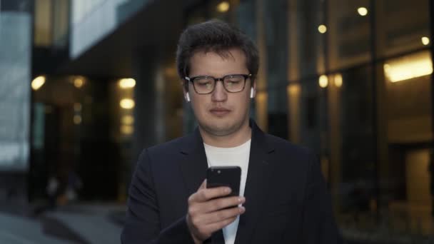 Homem rolando o telefone à noite em pé no fundo de janelas de vidro — Vídeo de Stock