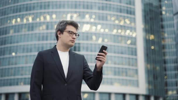 Мужчина в костюме стоит с телефоном на фоне бизнес-центра — стоковое видео