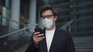 İş adamı koruyucu bir maske takıyor ve iş merkezinin yakınında bir telefon tutuyor.