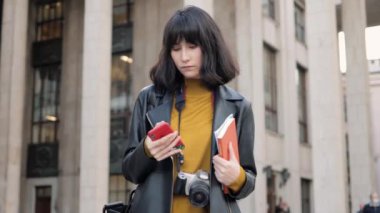 Fotoğraf makinesi olan bir kadın büyük binanın arka planında akıllı telefon kullanıyor.