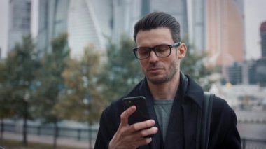 Siyah ceketli bir adam gökdelenlerin ve ağaçların arka planında telefonu kaydırıyor.