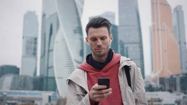 Hombre con capucha roja desplazando el teléfono en el fondo de los rascacielos — Vídeo de stock