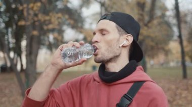 Kırmızı kapüşonlu adam parkta bir yudum su içiyor, ağır çekimde.