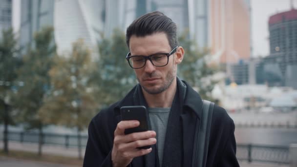 Людина в чорному пальто прокручує телефон на тлі хмарочосів і дерев — стокове відео