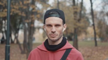 Kulaklıklı ve kırmızı kapüşonlu bir adam, şehir parkı ağaçlarının arka planında siyah bir şapka.