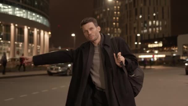 Zakenman in zwarte jas die 's nachts een taxi neemt, bedrijfsgebouwen — Stockvideo