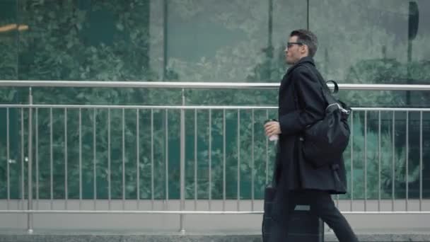 Mann in schwarzem Mantel mit Koffer läuft vor Glasfenster — Stockvideo
