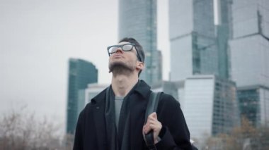 Siyah ceketli bir iş adamı gökdelenlerin arka planında gökyüzüne bakıyor.