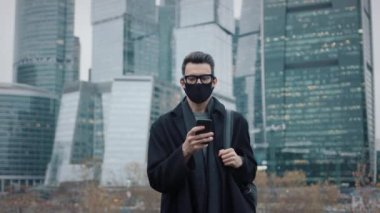 Koruyucu maskeli bir adam iş dünyasının arka planında ceketli bir gökdelende yürüyor.