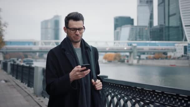 Mężczyzna w okularach z telefonem chodzącym na tle biurowych drapaczy chmur — Wideo stockowe