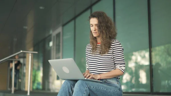 Jonge geconcentreerde vrouw krullend haar met laptop in gestripte trui, glazen kantoorraam — Stockfoto