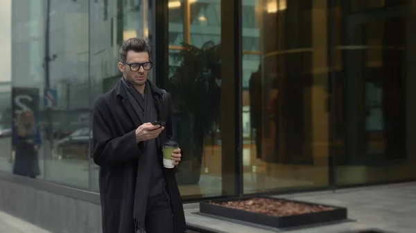 Hombre de negro y gafas con teléfono caminando por la calle con café — Foto de Stock