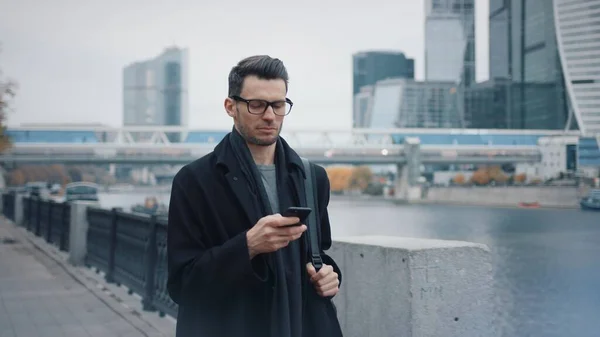 Hombre en gafas con teléfono caminando sobre fondo de rascacielos de negocios — Foto de Stock