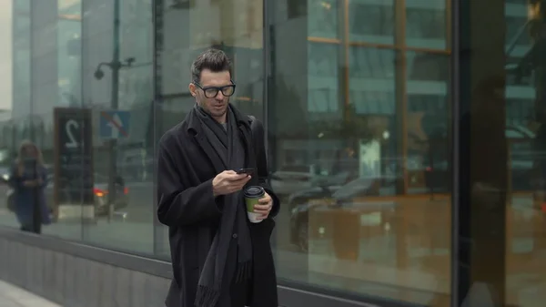 Hombre de negro y gafas con teléfono caminando por la calle con café — Foto de Stock