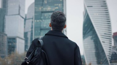 Palto giymiş bir adam iş binalarına doğru yürüyor, yavaş çekim gökdelenlere doğru.