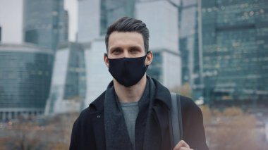 Gökdelenlerin arka planında koruyucu maskeli ve siyah ceketli bir iş adamı