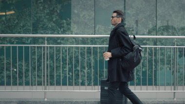 Siyah ceketli ve bavullu bir adam cam pencerelerde yürüyor.