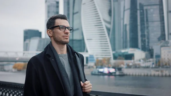 Homem de óculos andando no fundo de arranha-céus de negócios em casaco preto — Fotografia de Stock