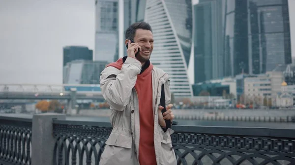 Hombre sonriendo, caminando con el teléfono en el fondo de los rascacielos de negocios — Foto de Stock