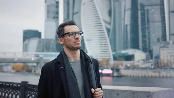 Uomo in occhiali che cammina sullo sfondo di grattacieli aziendali in cappotto nero — Foto Stock