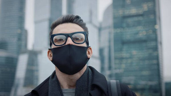 Man in eyeglasses and protective mask on background of business skyscrapers, looking up to the sky. Gimbal pan shot of businessman in coat standing near international business centre