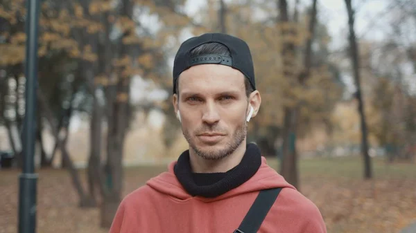 Hombre en auriculares y sudadera con capucha roja, gorra negra en el fondo de los árboles del parque de la ciudad — Foto de Stock