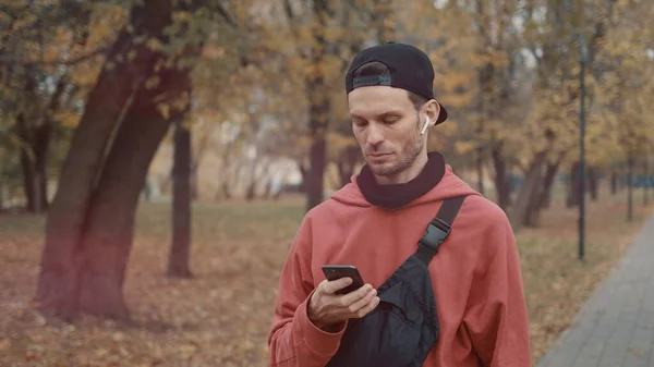 Homem de fones de ouvido e capuz vermelho, boné preto com telefone no parque da cidade — Fotografia de Stock