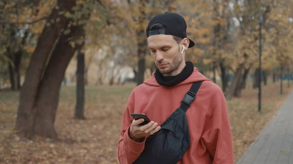 Homem de fones de ouvido e capuz vermelho, boné preto com telefone no parque da cidade — Fotografia de Stock