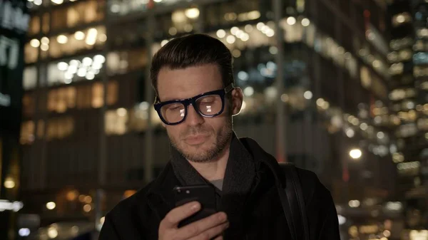 Homem de óculos com telefone no fundo do prédio de negócios à noite — Fotografia de Stock