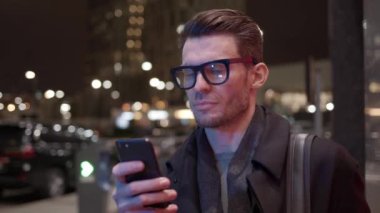 Gece vakti iş binasının arka planında telefonu olan gözlüklü adam.