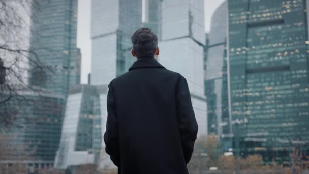 Férfi kabátban sétál felé üzleti épületek felhőkarcolók, lassított felvétel — Stock videók