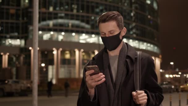 Homem em máscara protetora com telefone no fundo de edifícios de vidro durante a noite — Vídeo de Stock