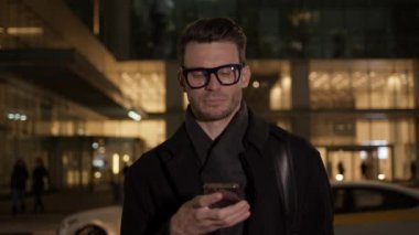 Gece vakti iş binasının arka planında telefonu olan gözlüklü adam.