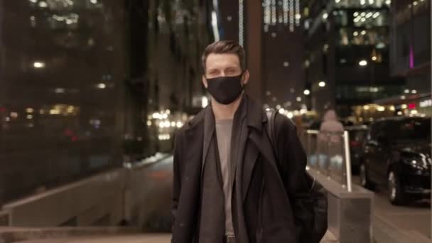 Bărbat în mască de protecție mergând pe fundalul clădirilor de sticlă în timpul nopții — Videoclip de stoc
