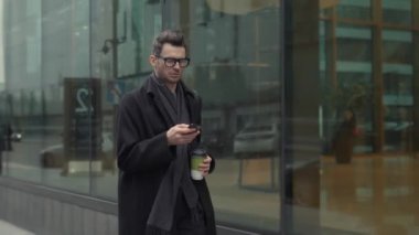 Siyahlı ve gözlüklü bir adam sokakta kahve içerken telefonla yürüyor.