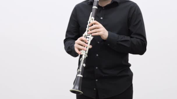 Человек в черной одежде играет на кларнете, стоя на белом фоне — стоковое видео