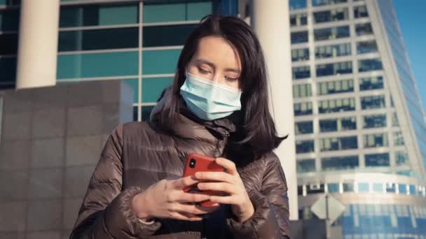 年轻的亚洲女人戴着防护面具，带着手机，沐浴在城市背景的阳光下 — 图库视频影像