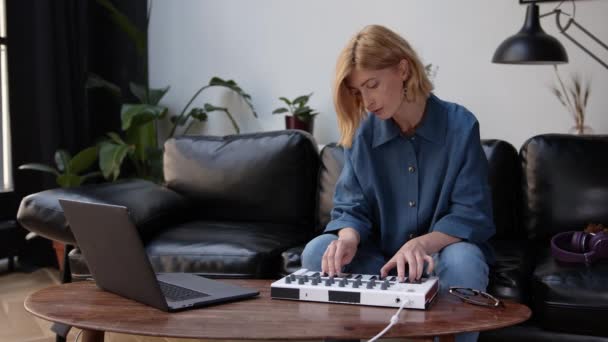 Женщина сидит на черном диване, играет на клавиатуре, ноутбук на кофейном столике — стоковое видео