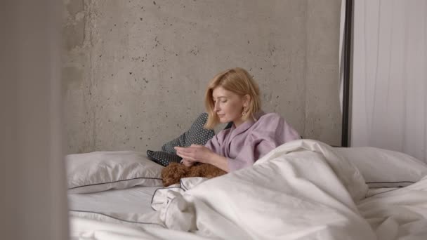 Jovem mulher na cama digitando no telefone, cão deitado em lençóis brancos — Vídeo de Stock