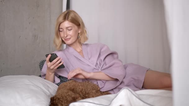 Ung kvinna sms:ar i telefon på sängen liggande med hund — Stockvideo
