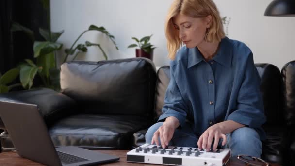 Femme assise sur un canapé noir enregistrant de la musique avec un ordinateur portable, jouant sur un clavier — Video